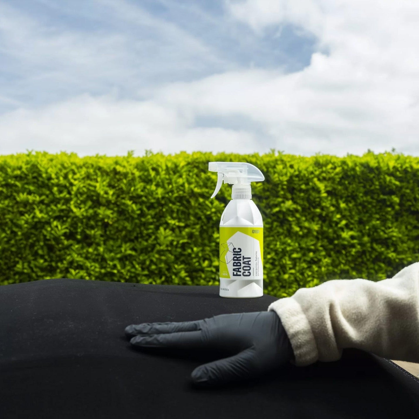Spray bottle labeled 'Gyeon Fabric Coat' held by a gloved hand against a green hedge and cloudy sky background.