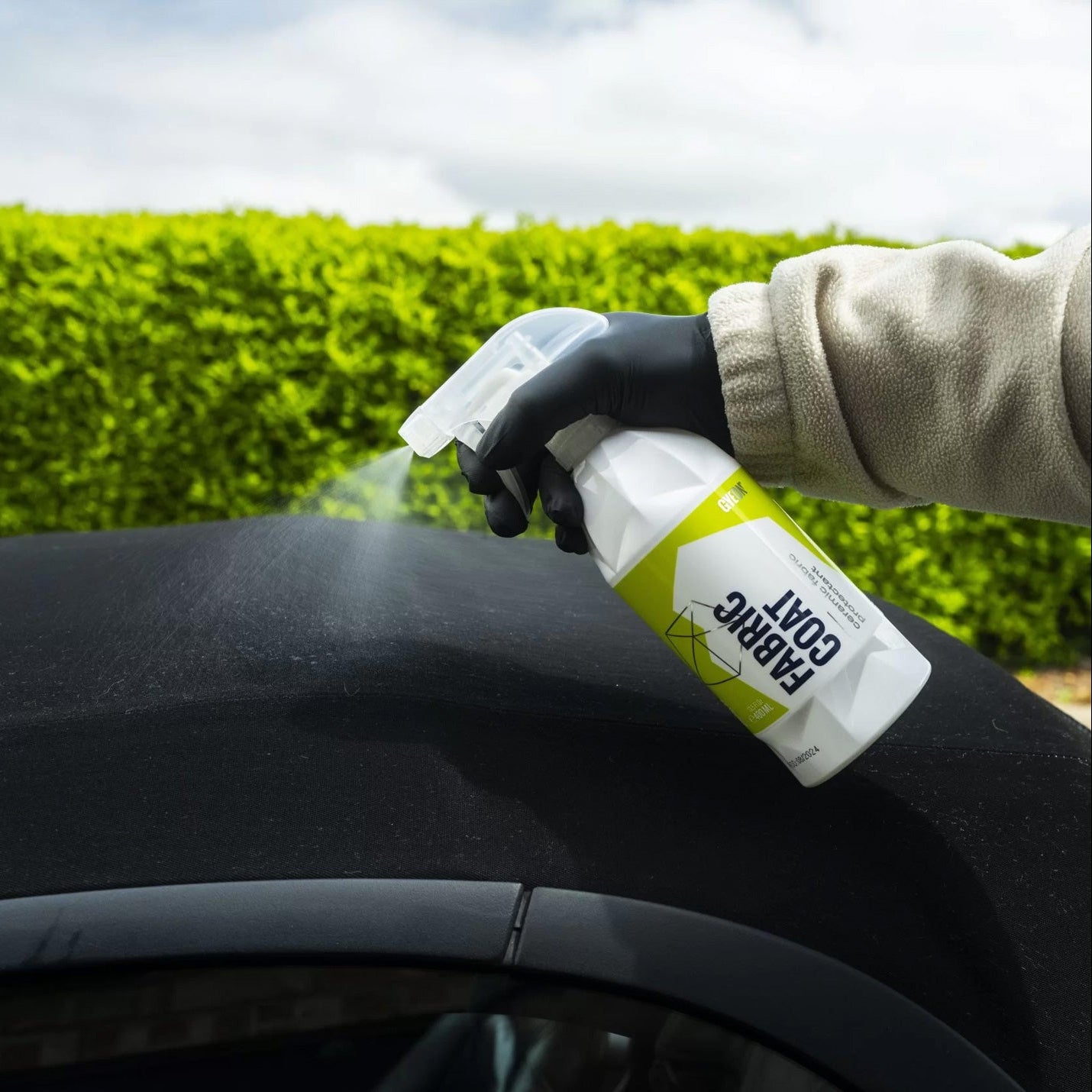 Person applying a Gyeon Fabric Coat to a car's surface with a green hedge in the background