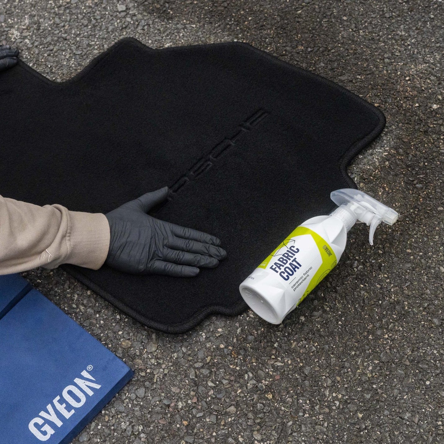 Person wearing gloves handling a black car mat with a bottle of Gyeon Fabric Coat on a concrete surface.