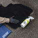 Person wearing gloves handling a black car mat with a bottle of Gyeon Fabric Coat on a concrete surface.