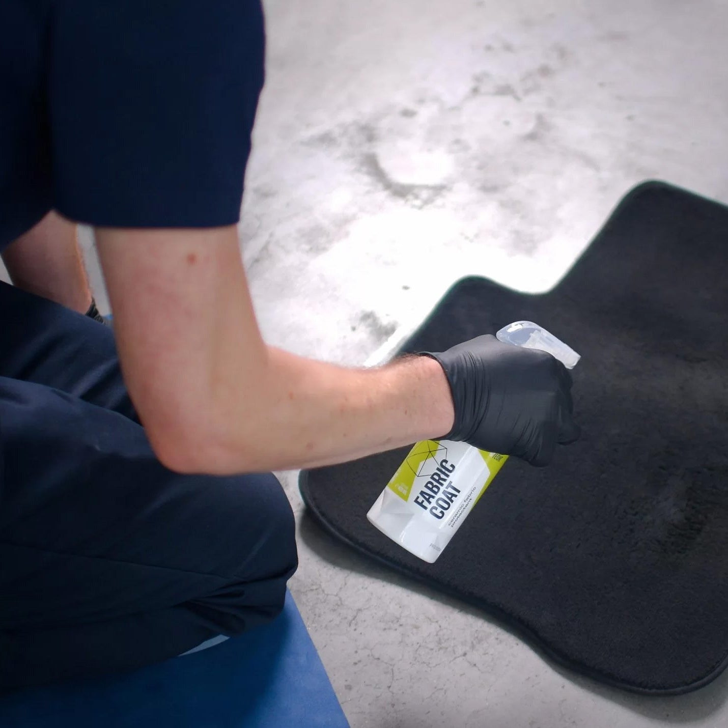 Person coating a black car mat with Gyeon Fabric Coat and a white cloth on a concrete surface.
