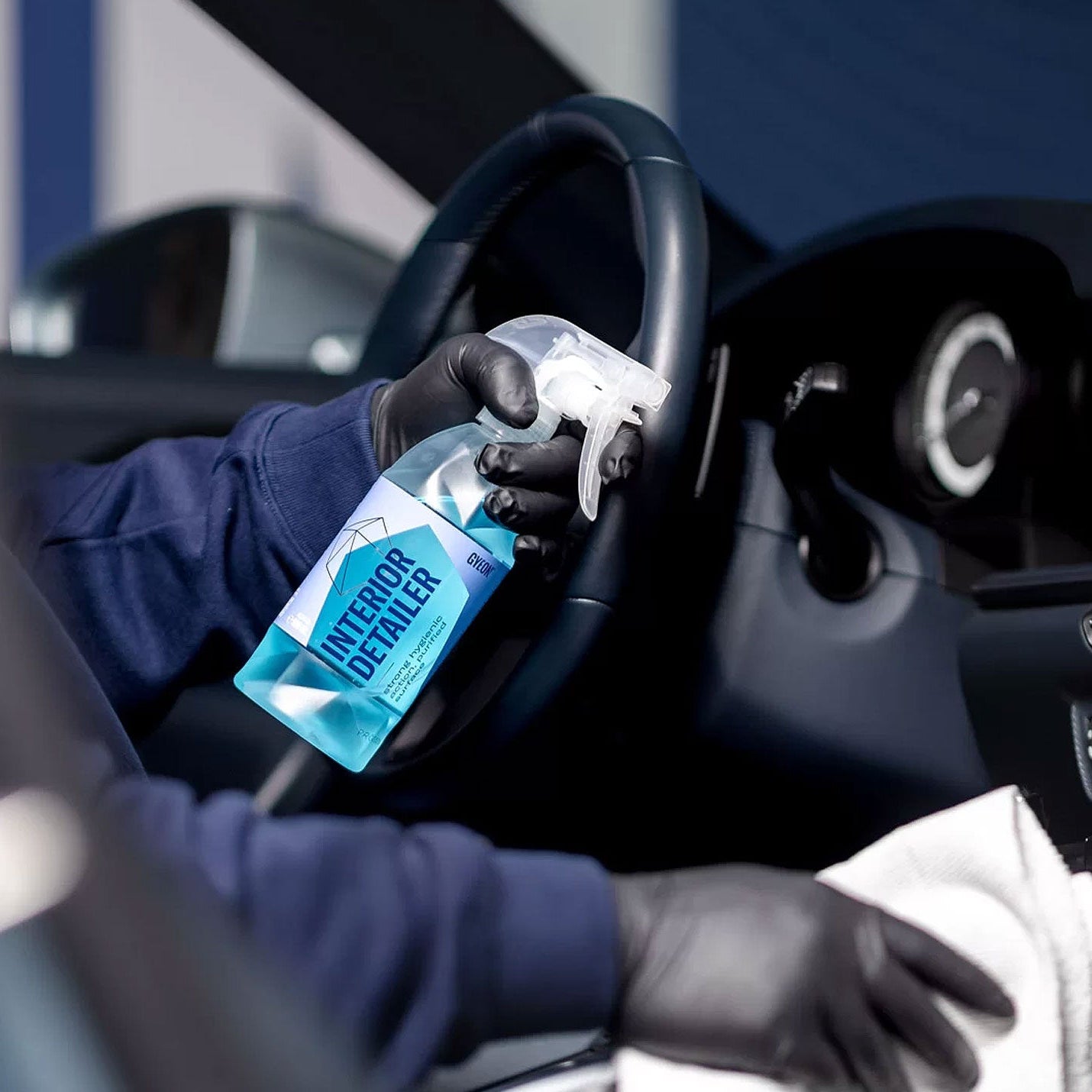 Person cleaning car interior with a spray bottle labeled 'Gyeon Interior Detailer' and a cloth.
