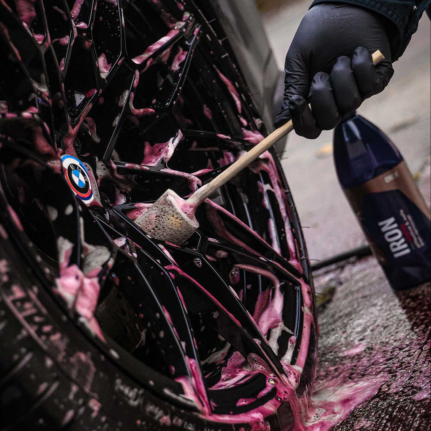 Person cleaning a car tire with pink soap and a brush, wearing black gloves.