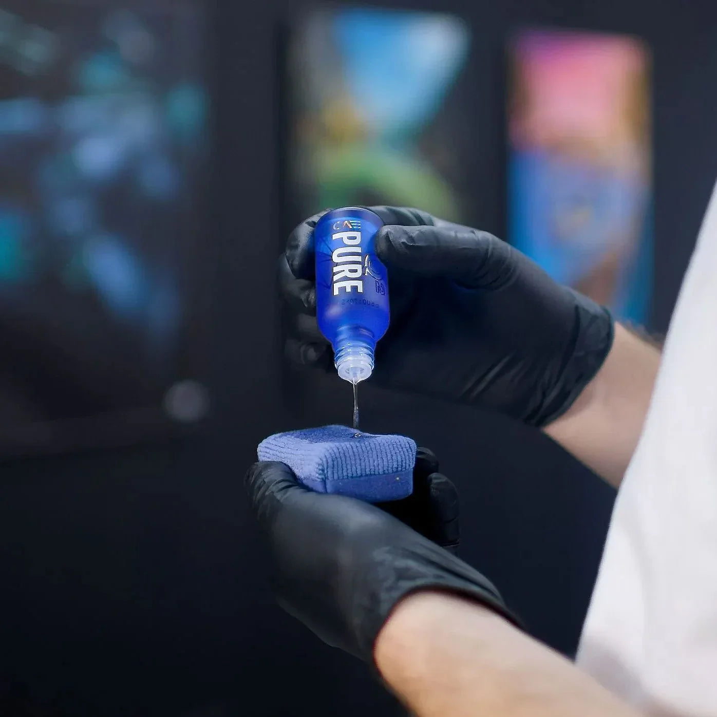 Person wearing black gloves applying a blue liquid from a bottle labeled 'PURE' onto a blue cloth against a blurred background.