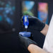 Person wearing black gloves applying a blue liquid from a bottle labeled 'PURE' onto a blue cloth against a blurred background.