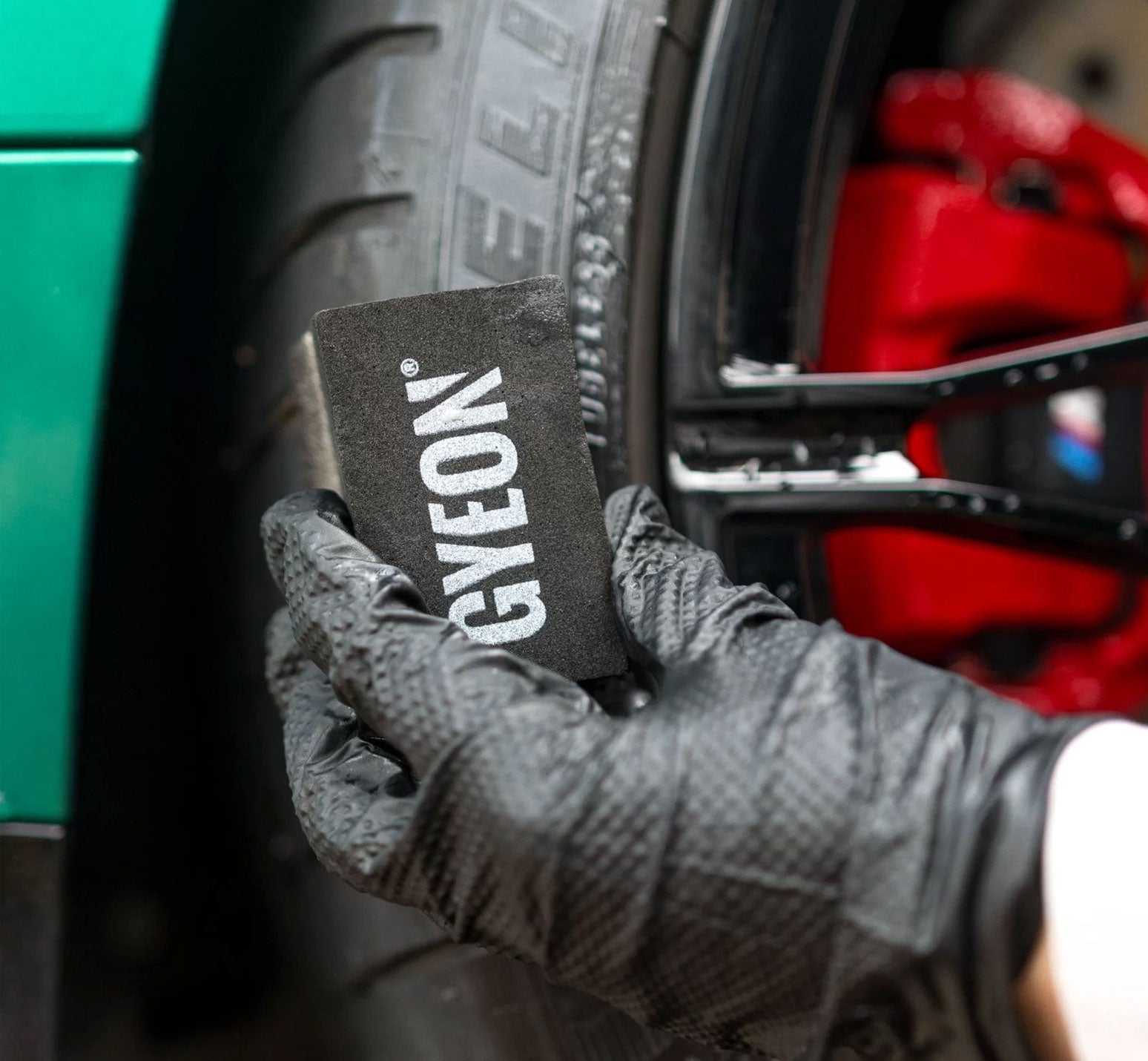 Hand wearing a grey glove holding a Gyeon tyre dressing applicator near a car tire and red brake caliper.