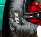 Hand wearing a grey glove holding a Gyeon tyre dressing applicator near a car tire and red brake caliper.