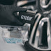 Gyeon tire applicator and packaging held by a gloved hand, with a blurred car wheel in the background.
