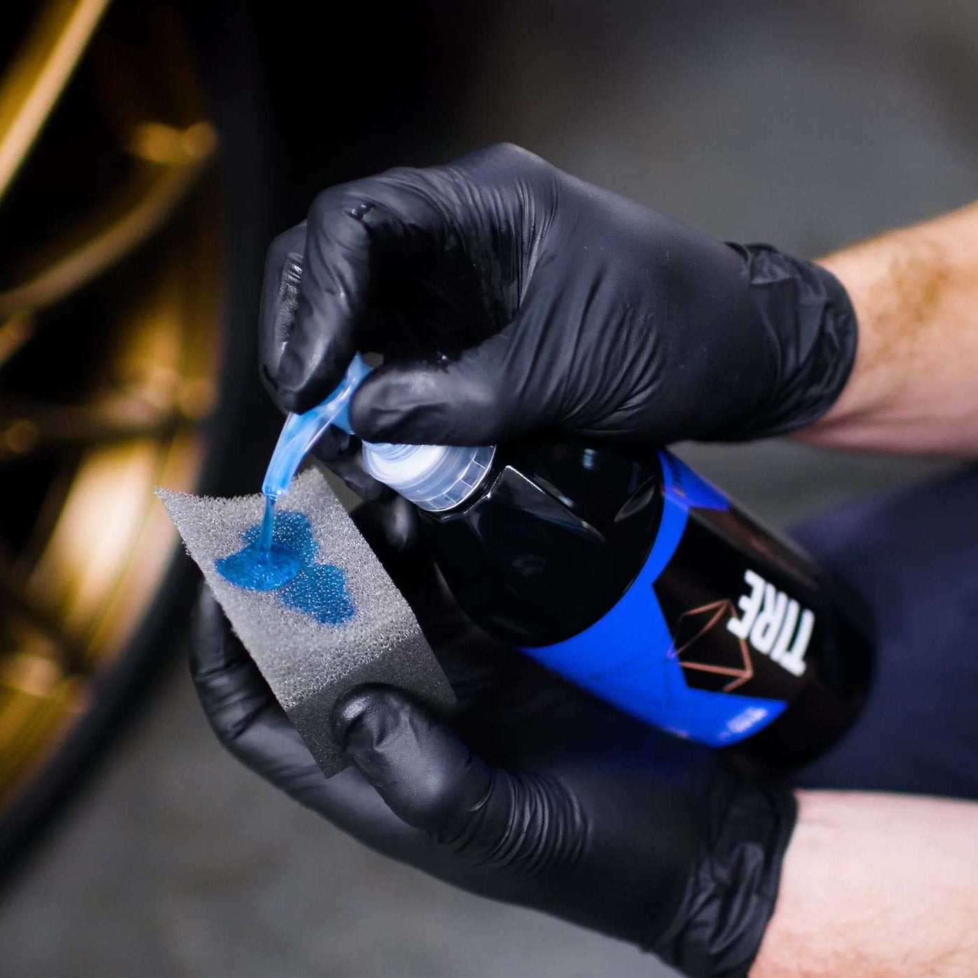 Person applying a blue substance from a bottle of Gyeon Tire Dressing onto a cloth, with a blurred background.