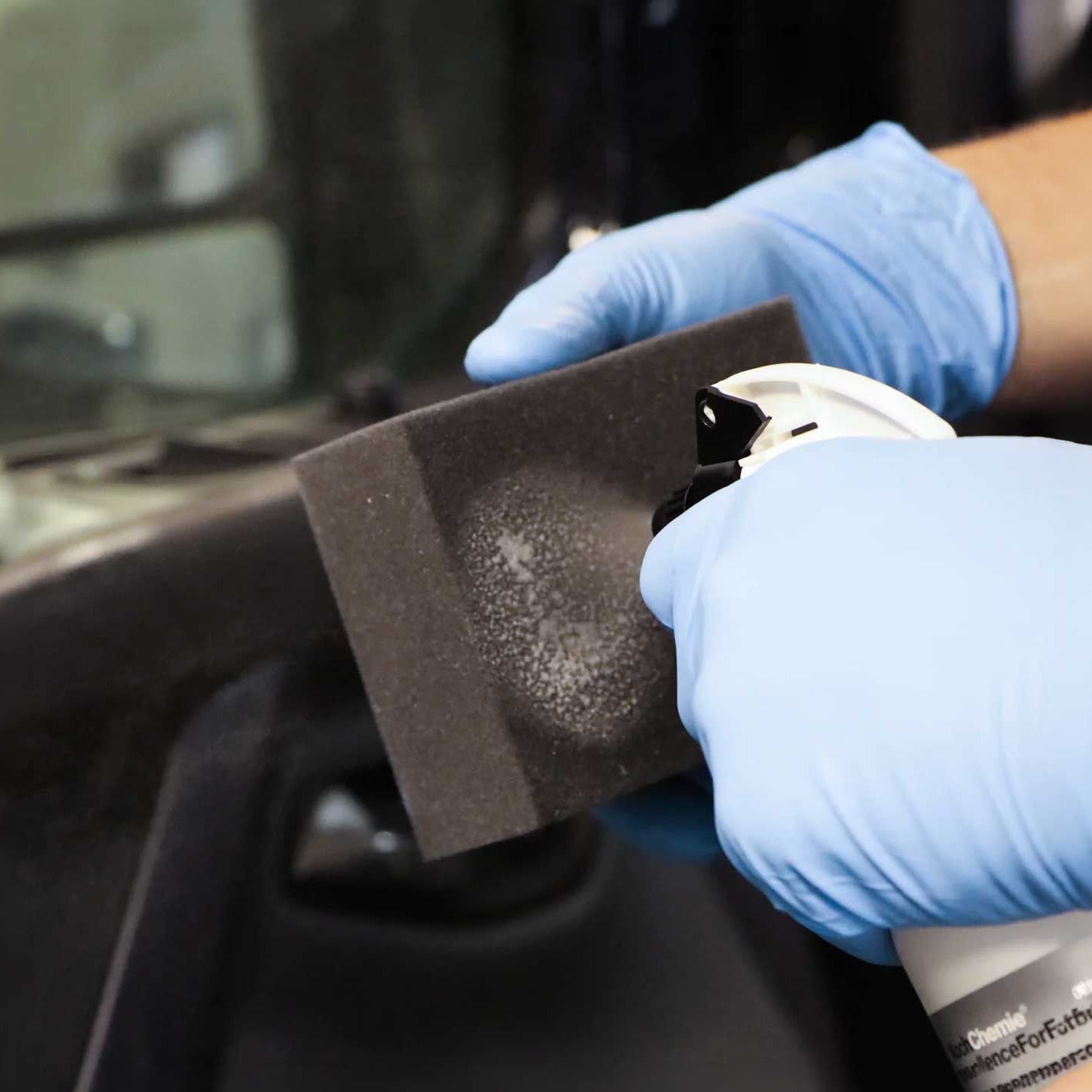 A close-up of a person wearing blue gloves holding a dark gray foam block and spraying a cleaning solution from a white spray bottle onto a dusty area.