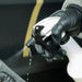 A close-up of a hand wearing a black nitrile glove holding a white spray bottle with a black trigger nozzle, spraying a fine mist. The background is a dark, out-of-focus surface with a yellow stripe.