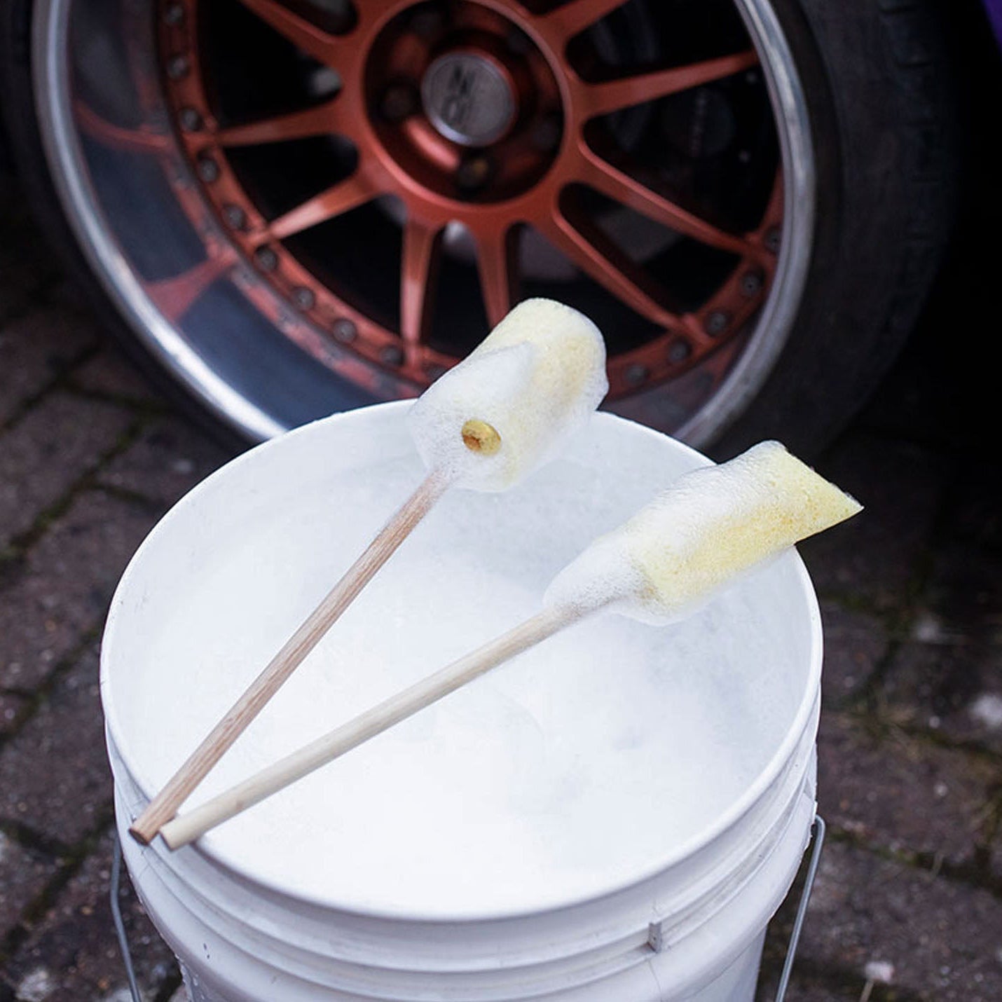 Two Sponge Guru Oval Wheel Brushes on a white bucket with a car wheel in the background