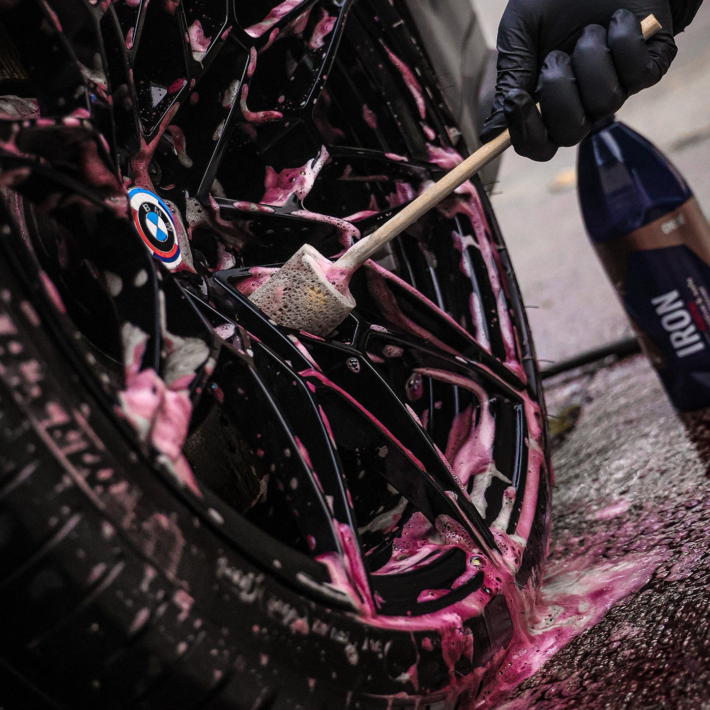 Person cleaning a car wheel with a Sponge Guru Wheel Brush pink soap suds using a brush and bottle labeled 'Gyeon Iron'.